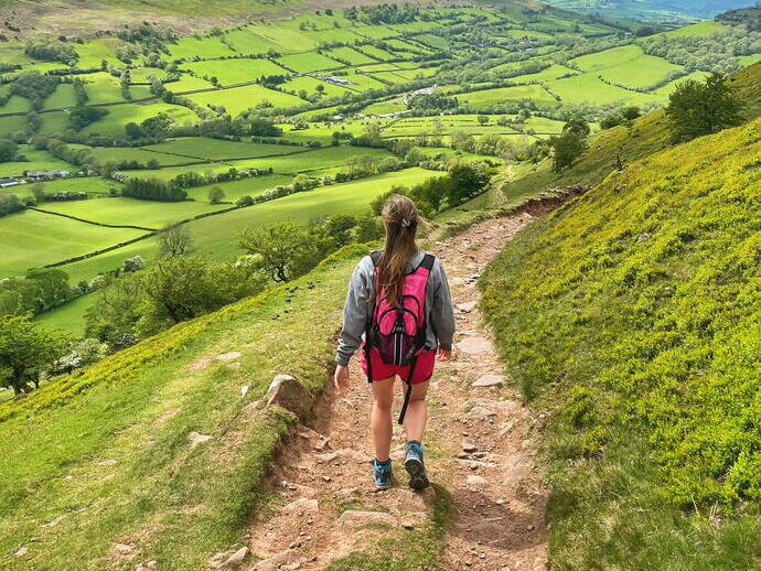 Rückansicht einer jungen Frau, die auf einem Weg durch ein üppig grünes Tal und Hügel spaziert