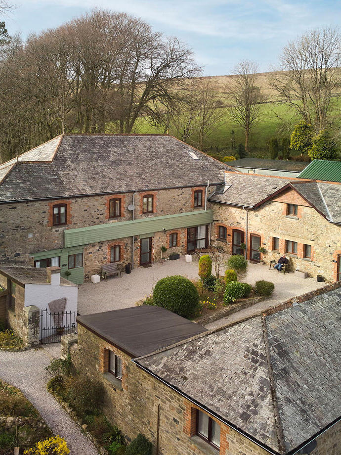 A number of brick buildings arranged around a gravel courtyard, surrounded by trees, hills and fields. The Cottages at Blackadon Farm - Bronze award winner for the Ethical, Responsible and Sustainable Tourism Award at the VisitEngland Awards for Excellence 2023.