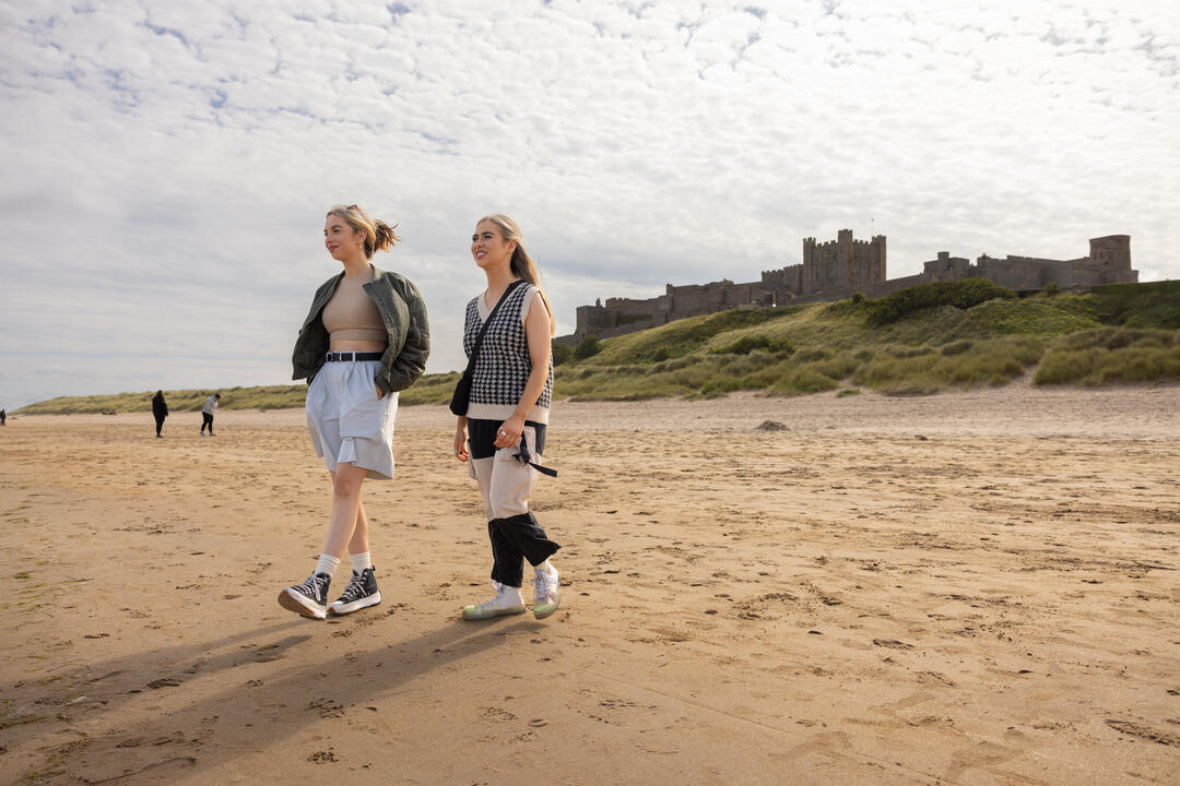 Two women walking in the sand dunes by the sea with a castle in the background