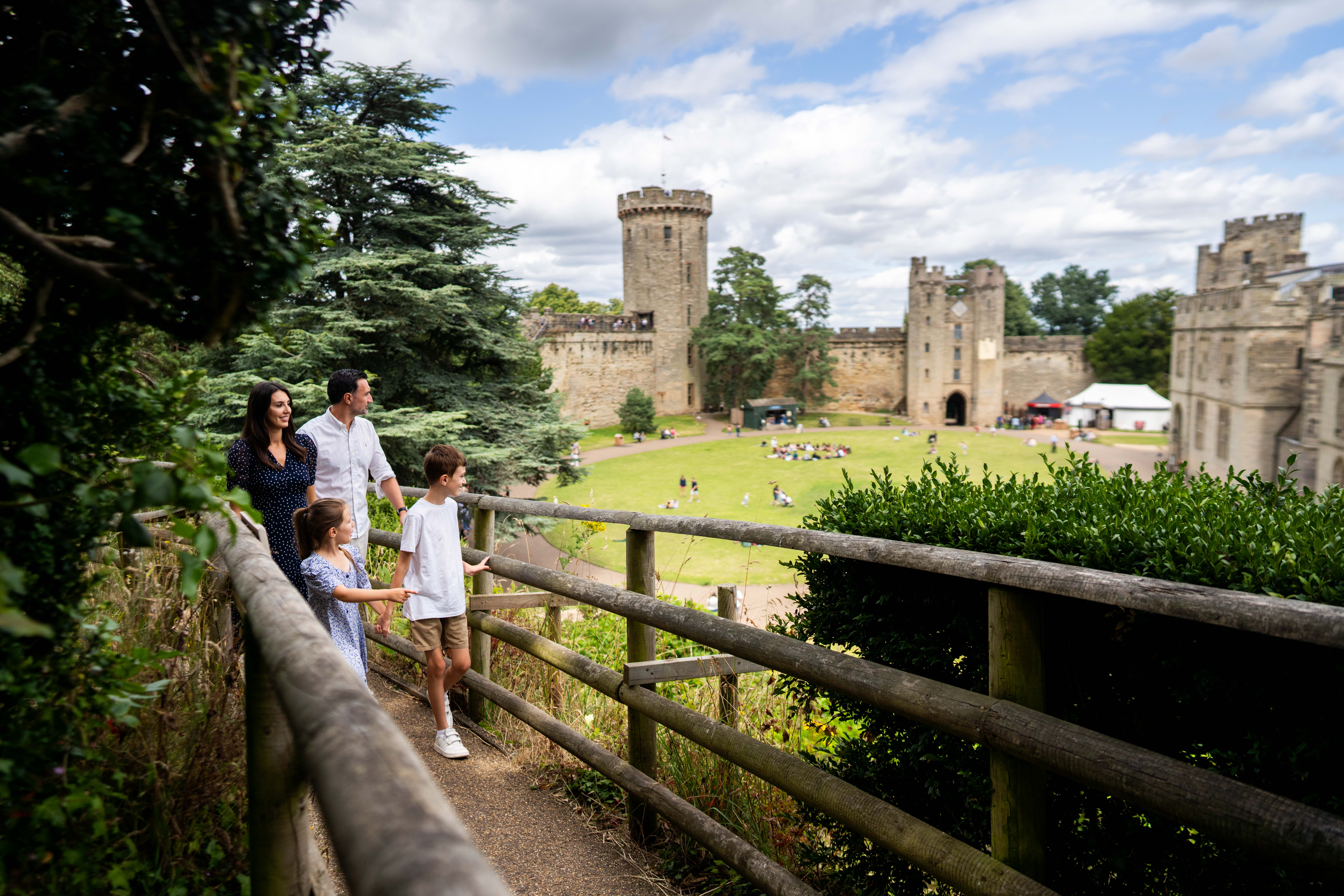 Familia caminando sobre un puente de madera con castillo de piedra y jardín verde al fondo.