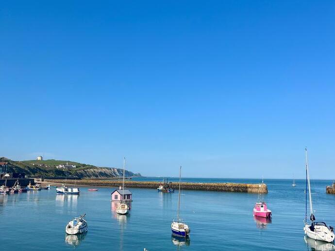 Small boats float in a calm harbor with a clear blue sky, coastal cliffs, and distant houses visible on shore.