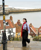 Person walking up stone steps by red-roofed houses overlooking a river and seaside harbor, with cliffs and coastline in the background.