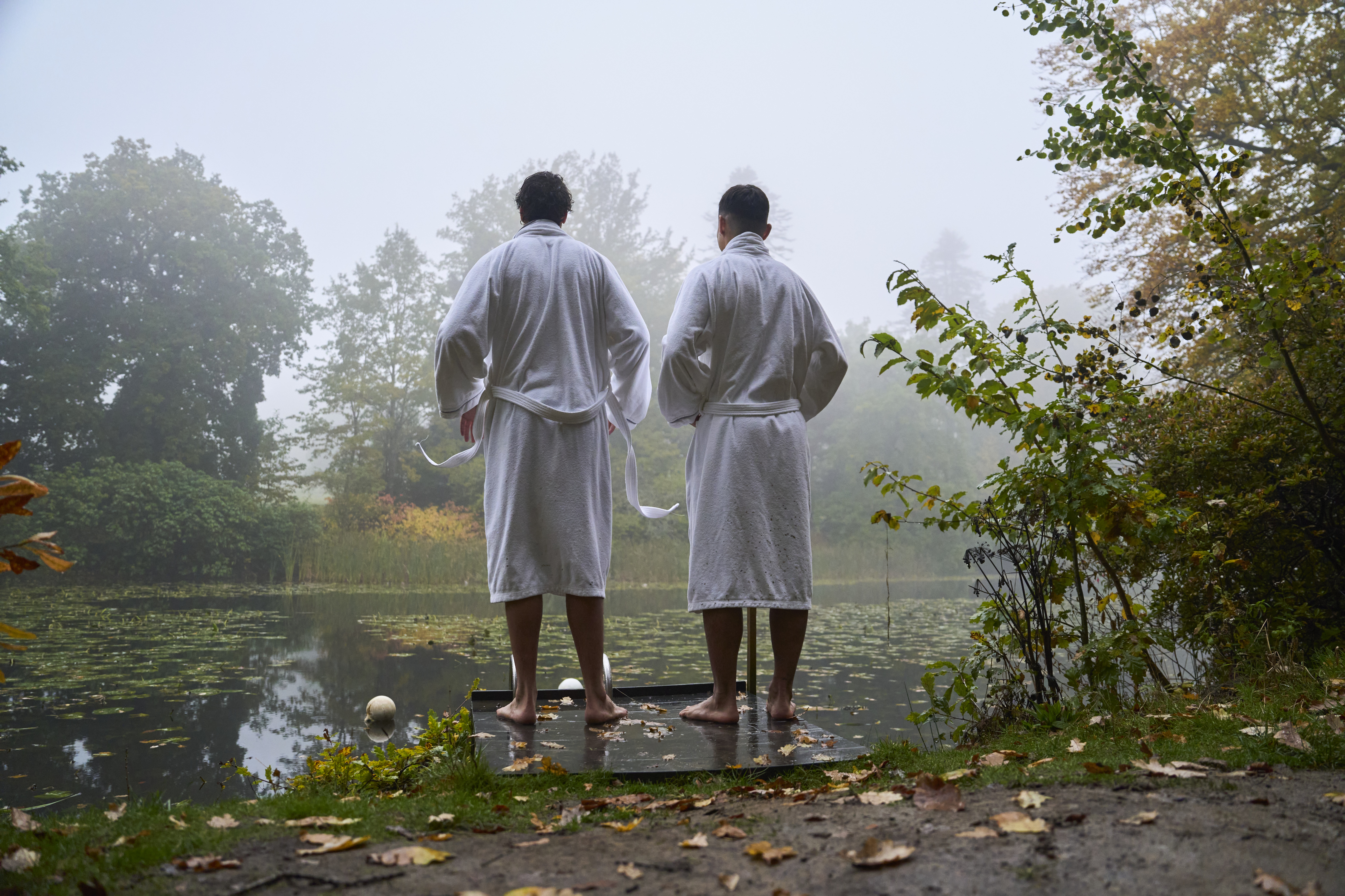 Two men in robes on the swimming platform of a lake in an autumnal setting