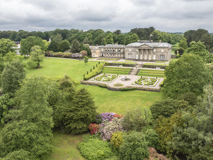 A stately neoclassical mansion with formal gardens, surrounded by lush parkland and dense trees under a cloudy sky.