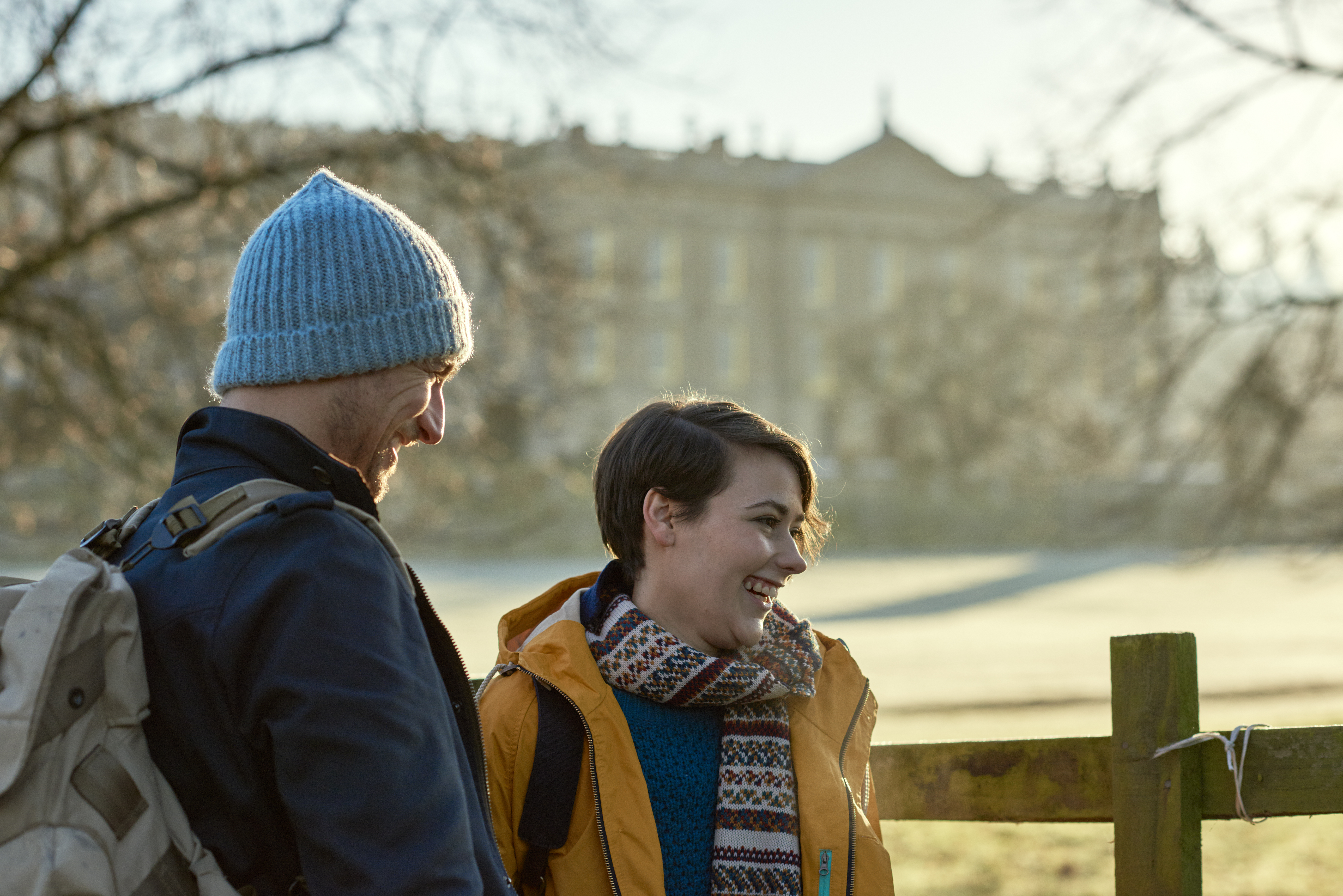 Pareja sonriendo y de pie bajo el sol invernal frente a una casa de campo