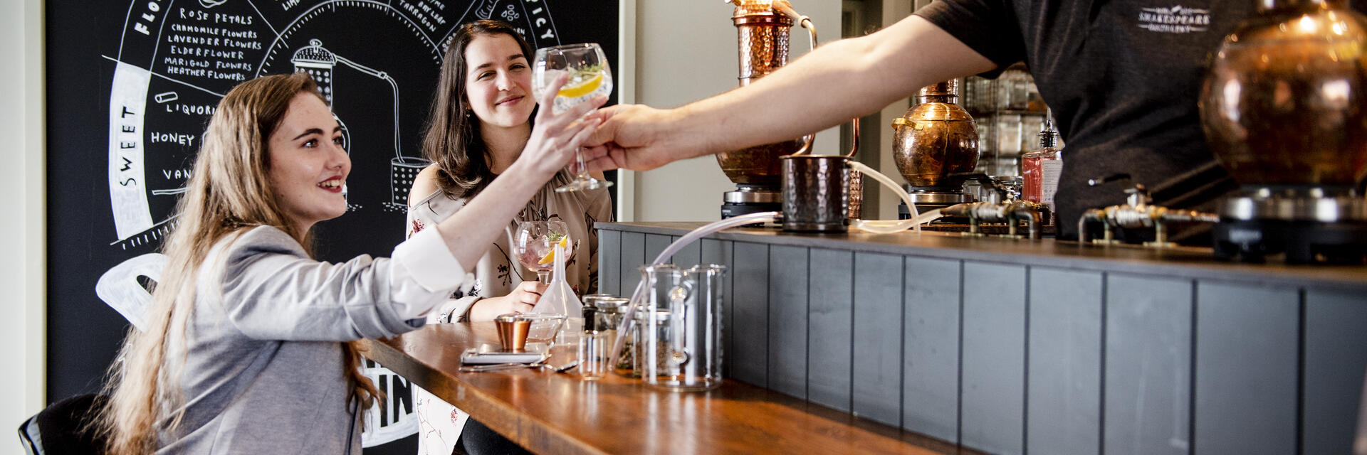 Zwei Frauen, eine davon im Rollstuhl, werden von einem Barkeeper in einer Brennerei-Bar mit einem Getränk bedient
