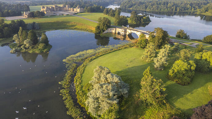 Vista aérea de un palacio del siglo XVIII, rodeado de jardines, árboles y un lago atravesado por un puente.
