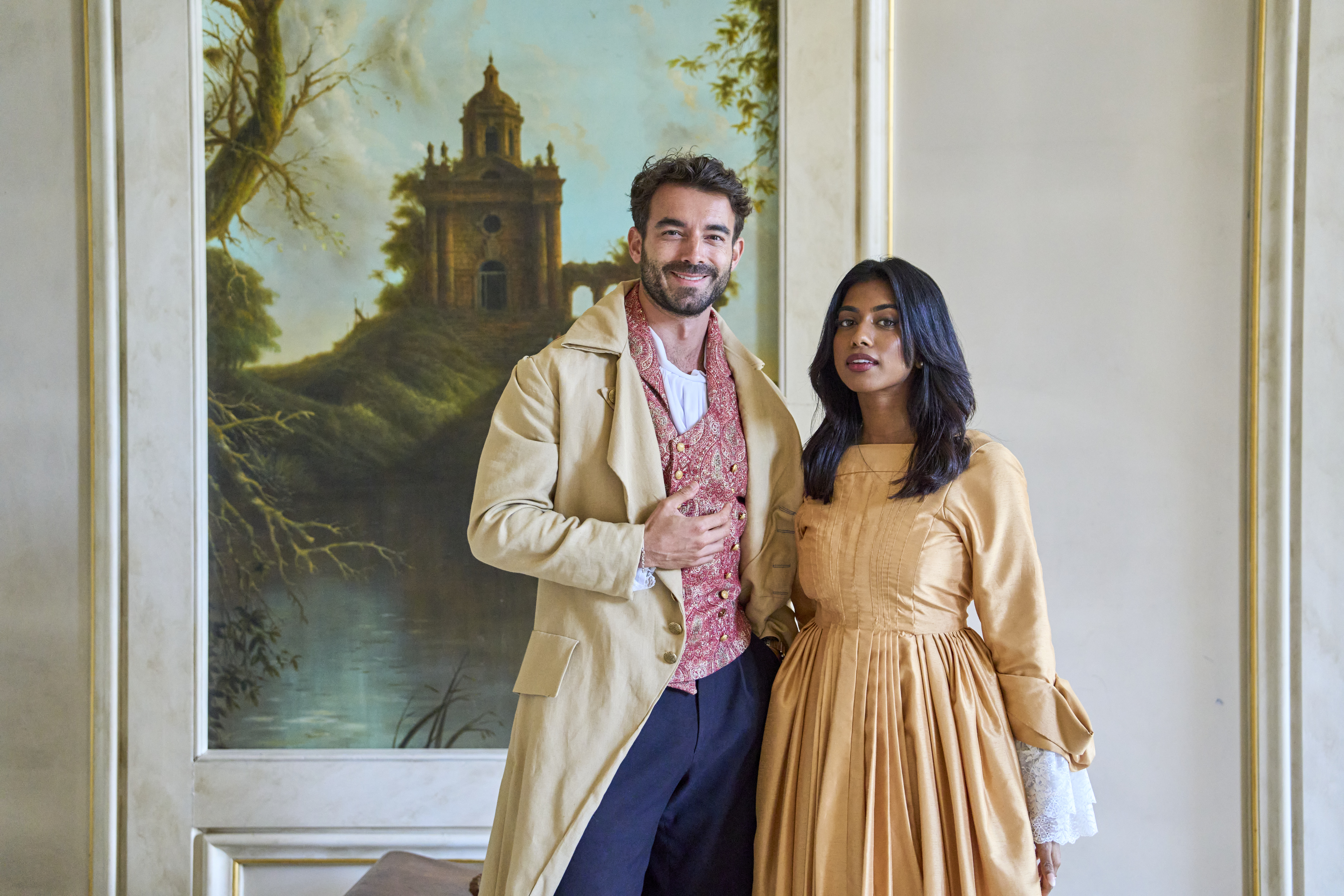 Ein Mann und eine Frau in historischen Kostümen stehen in einem dekorierten Raum vor einem Gemälde mit Landschaft und altem Gebäude.