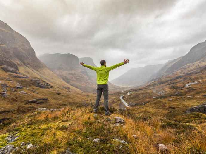 Rückansicht eines Mannes mit ausgestreckten Armen, der in einem Tal steht und auf die in Nebel gehüllten Berge blickt
