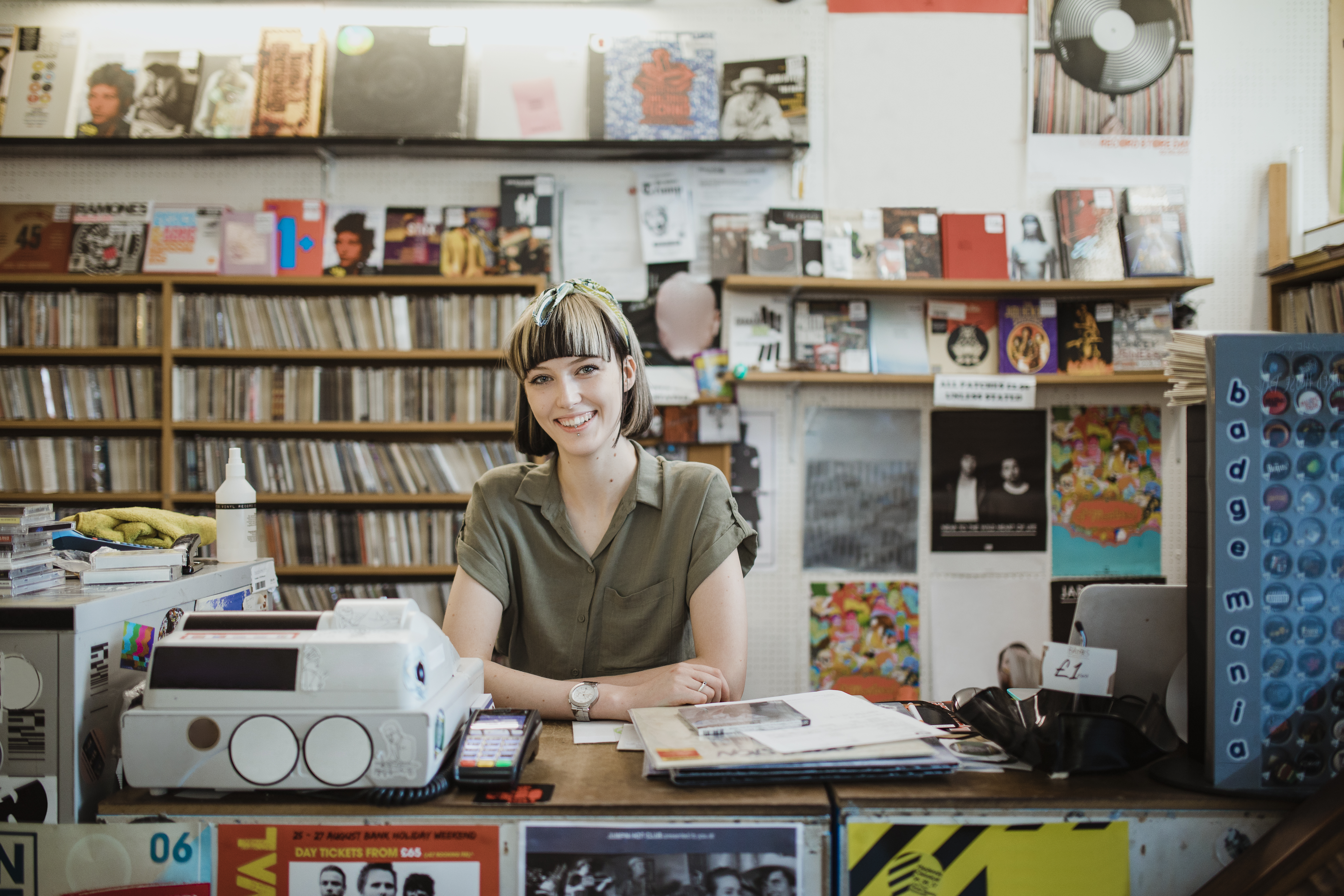 Young woman smiling, behind a counter in a record shop, with the till and card reader in front of her