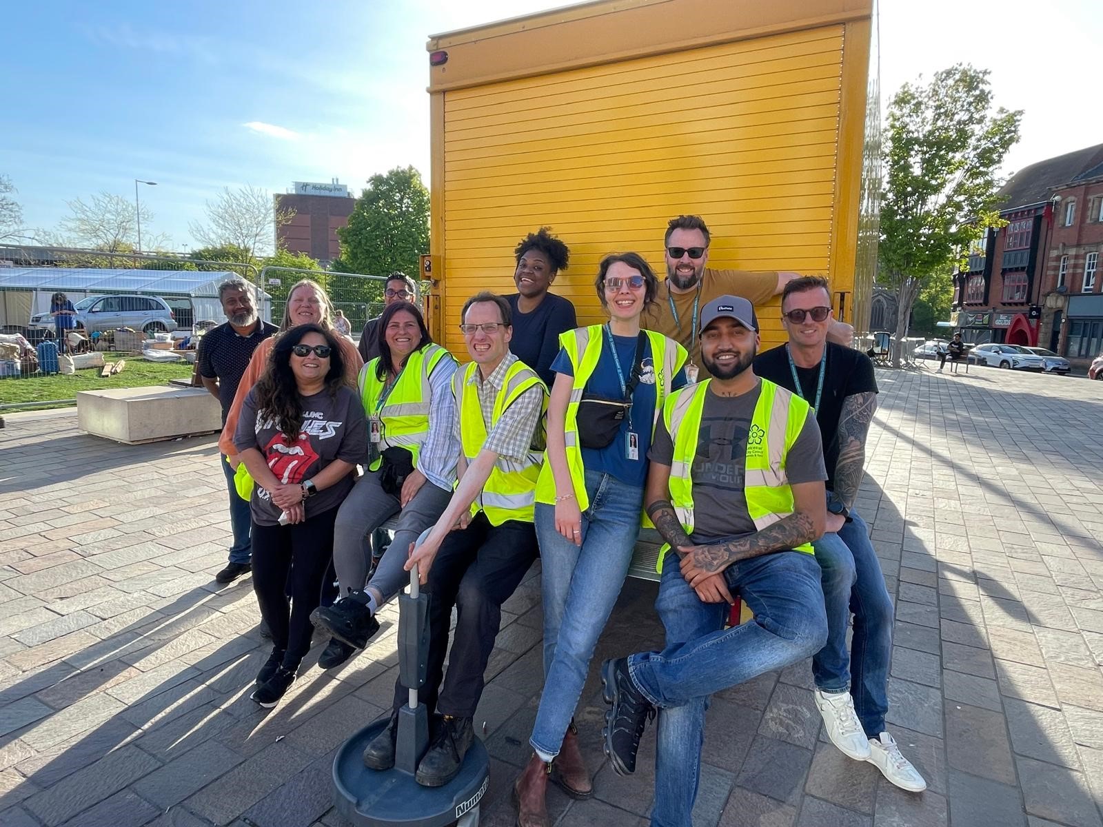 A group of people wearing high-visibility vests and casual clothes pose and smile together outdoors in front of a yellow truck on a sunny day.