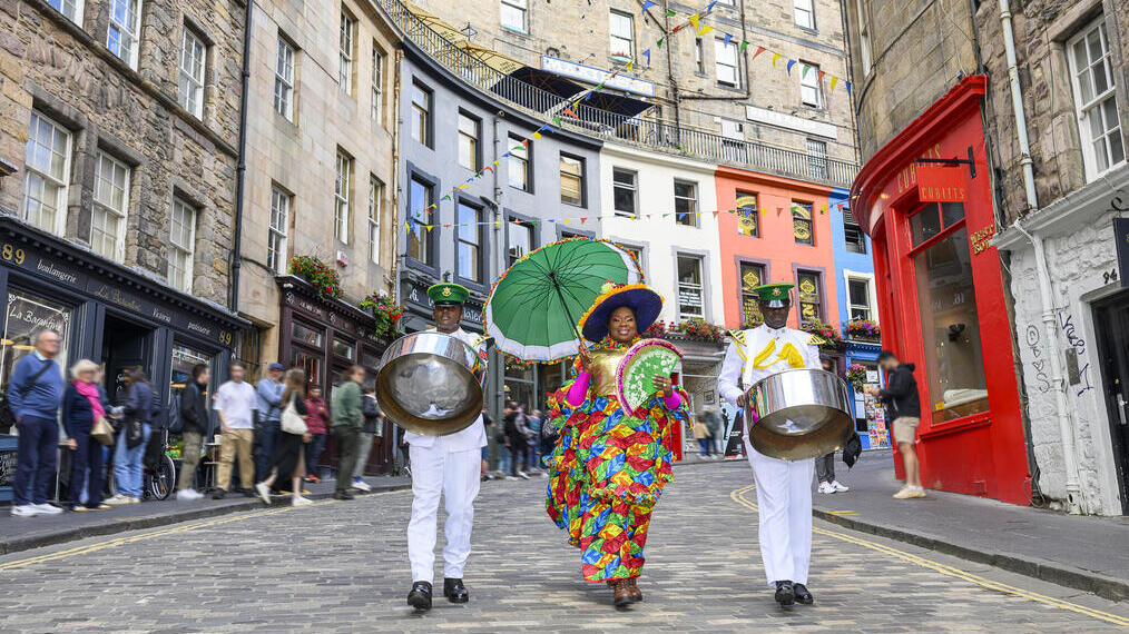 Festival performers in traditional dress walking down a street carrying steel drums and a parasol