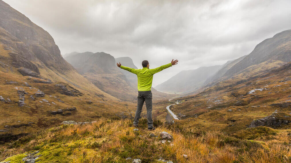 Rückansicht eines Mannes mit ausgestreckten Armen, der in einem Tal steht und auf die in Nebel gehüllten Berge blickt