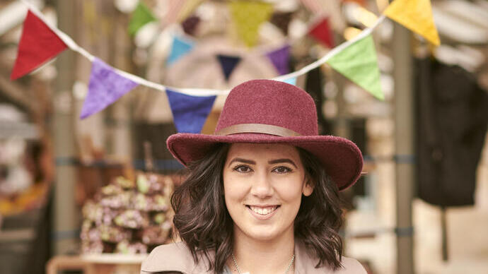 Person in a maroon hat smiles while holding dessert at a market with colorful bunting and wooden crates.
