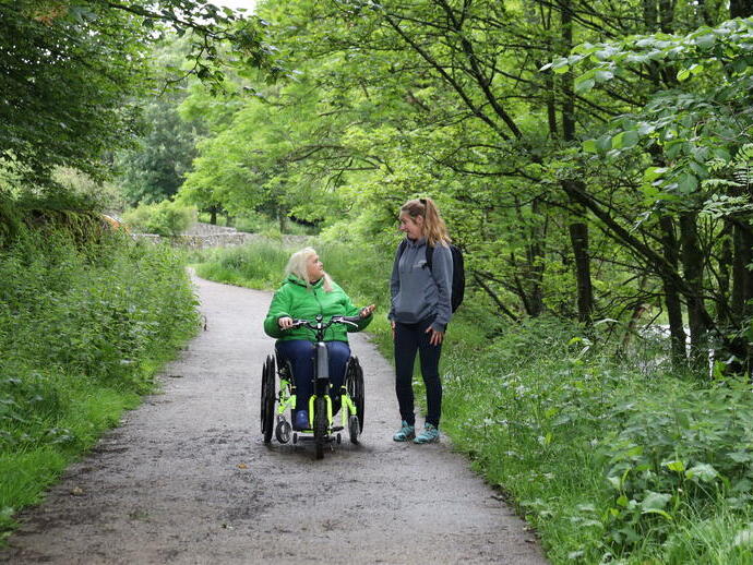 Dos mujeres, una en una bicicleta con silla de ruedas, en un camino rodeado de árboles
