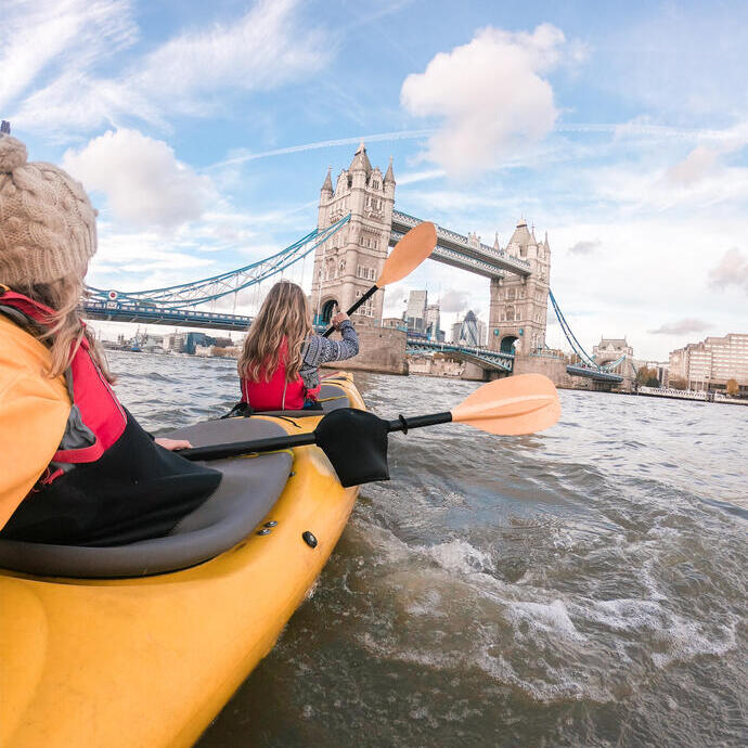 Dos mujeres remando en kayak por un río hacia un puente en una ciudad