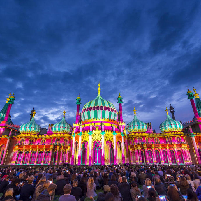 Crowd sitting on the ground in front of a large ornate building lit up with bright colours