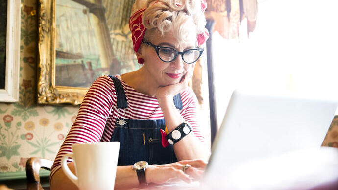Close up of a blond woman wearing a stripy top and red head scarf, having a coffee and looking down at a lap top screen, smiling.