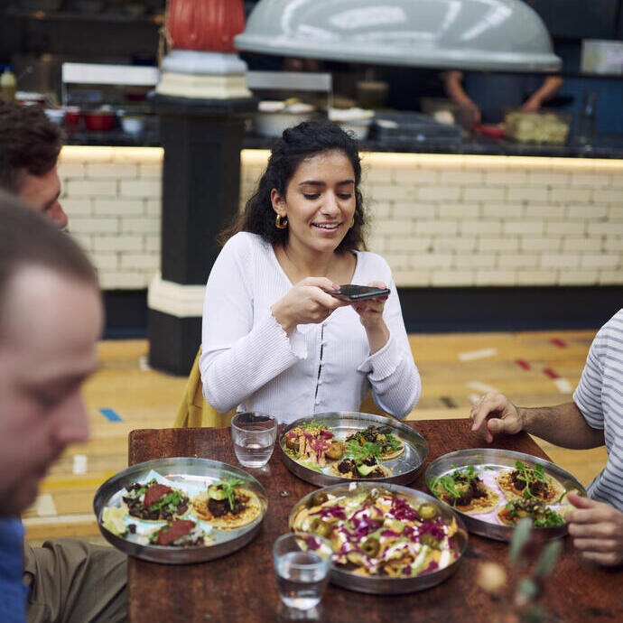 Mujer sentada con amigos en una mesa, tomando una foto de la comida en un patio de comidas