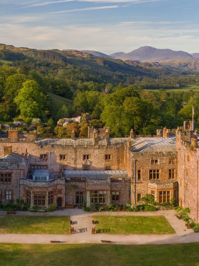 A country house with decorative mock foundations against a backdrop of tree covered hills, including Scafell Pike. Muncaster Castle - Silver award winner for Small Visitor Attraction of the Year at the VisitEngland Awards for Excellence 2023.