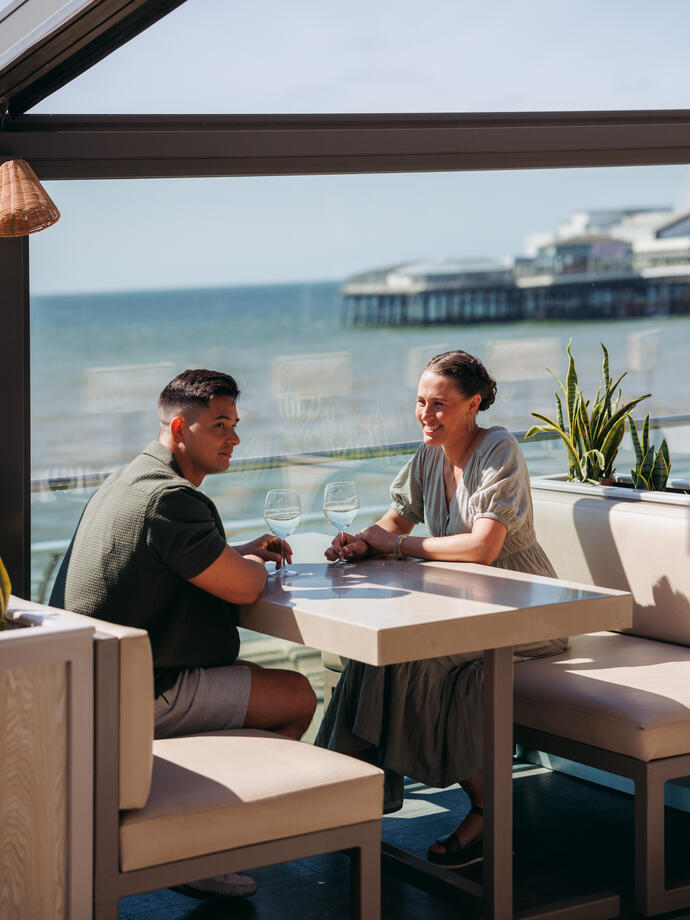 A couple sitting in an enclosed beachside restaurant overlooking the ocean and a pier.