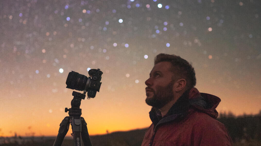 A person in a red jacket stands next to a camera on a tripod under a starry night sky, with a glowing orange horizon in the background.