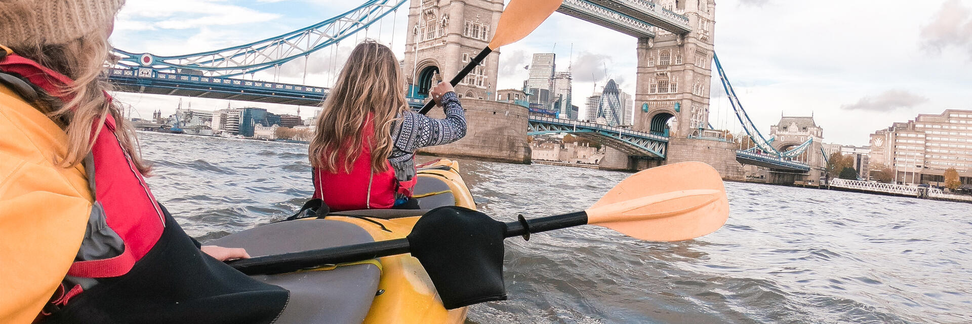 Two women paddling in a kayak on a river towards a bridge in a city