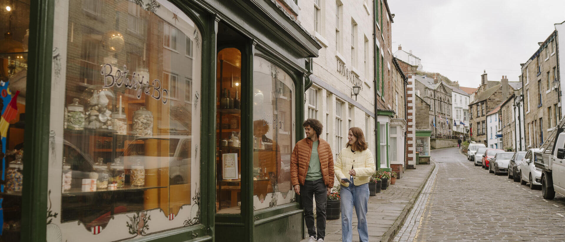 Two people walk past a vintage sweet shop on a cobbled street lined with historic buildings and parked cars on a cloudy day.