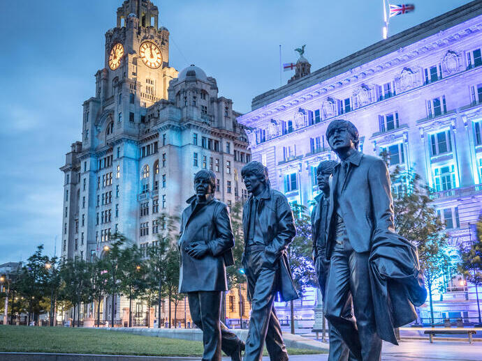 A sculpture of four men with a large building with clock tower behind, lit up in the evening.
