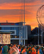 A crowd of people at a festival watching acrobats performing in the air in front of Symphony Hall, Birmingham at sunset
