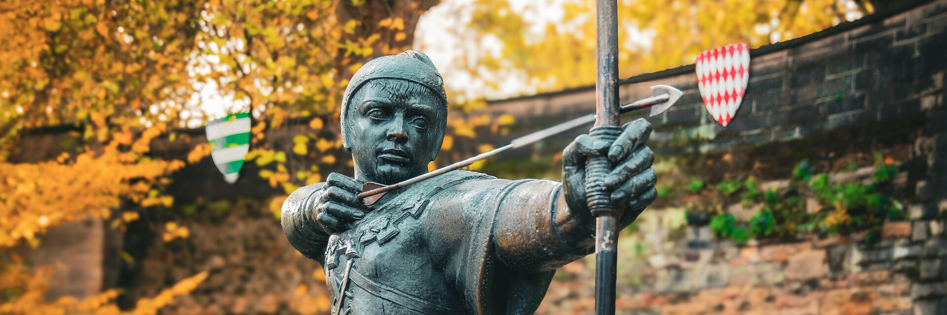 Statue of an archer drawing a bow, set outdoors with autumn trees and a stone wall in the background.