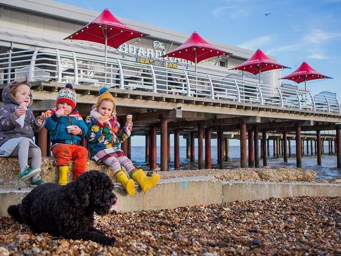 Three children eat ice cream near a pebble beach, with a black dog in front and a pier with red umbrellas in the background.
