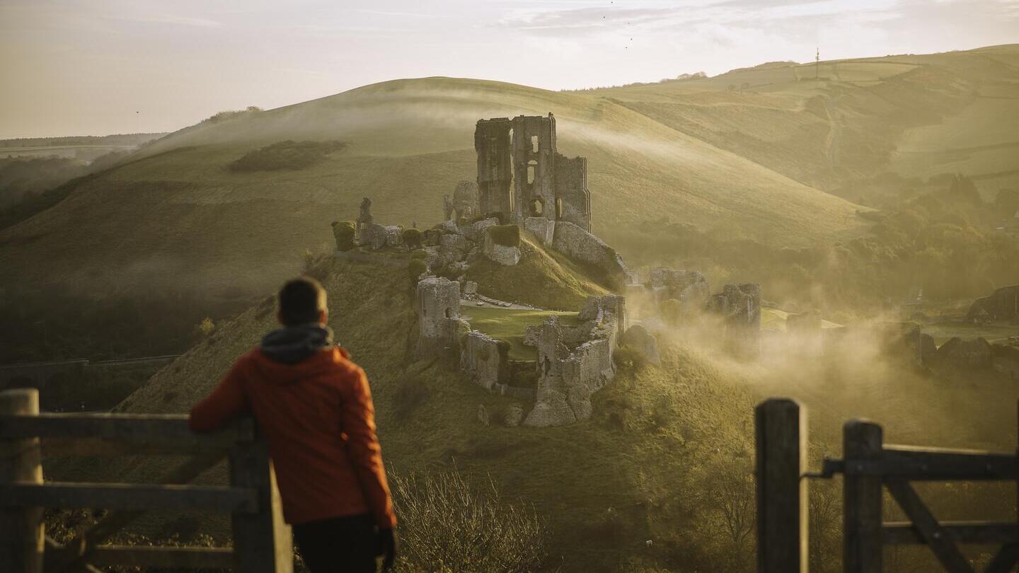 Uomo appoggiato al palo di un cancello, che guarda le rovine di un castello