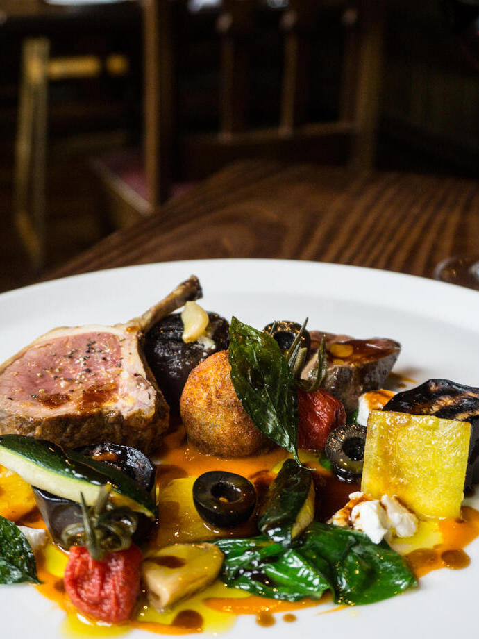 A white plate of colourful food including lamb and mediterranean vegetables, with a glass of red wine in the background 