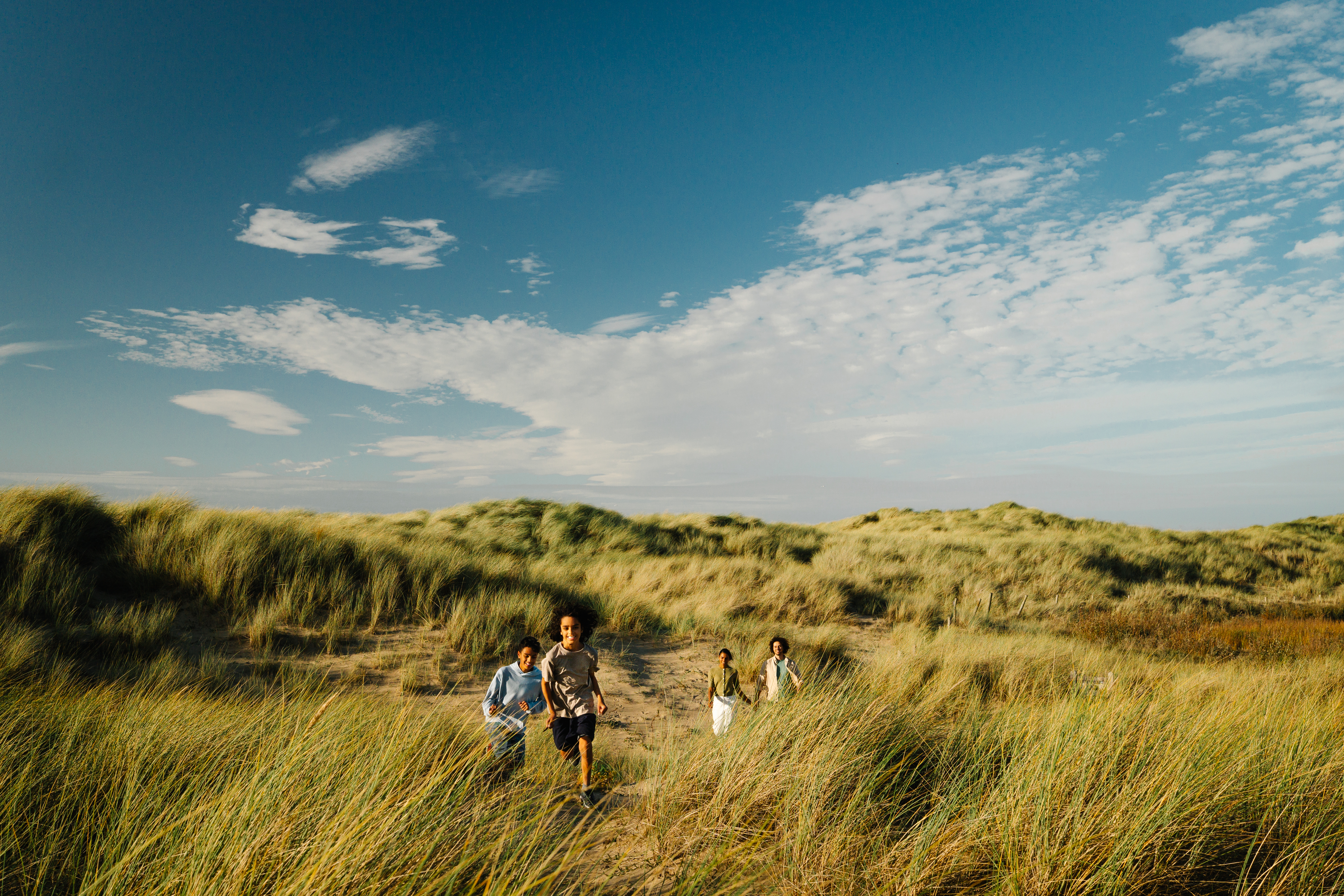 Children running through grassy sand dunes under a bright blue sky with scattered clouds.