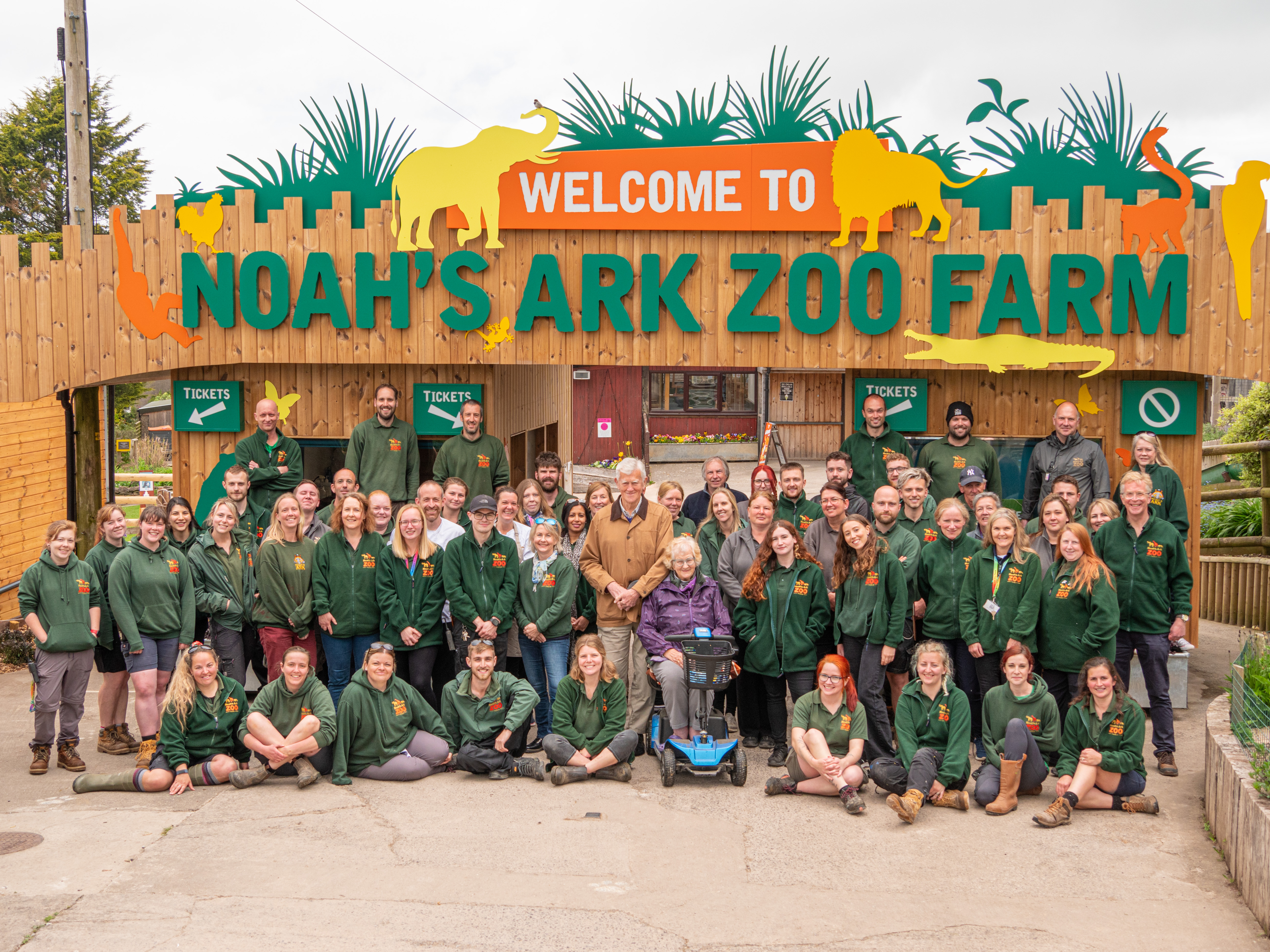A sign which reads "Welcome to Noah's Ark Zoo Farm" with various staff members standing in front wearing matching branded tops. Noahs Ark Zoo Farm - Gold award winner for the Accessible and Inclusive Tourism Award at the VisitEngland Awards for Excellence 2023.
