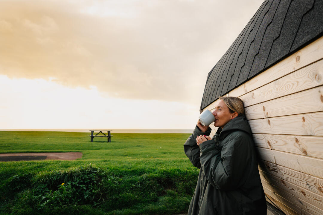 A woman drinks a hot drink while leaning against a sauna by the sea