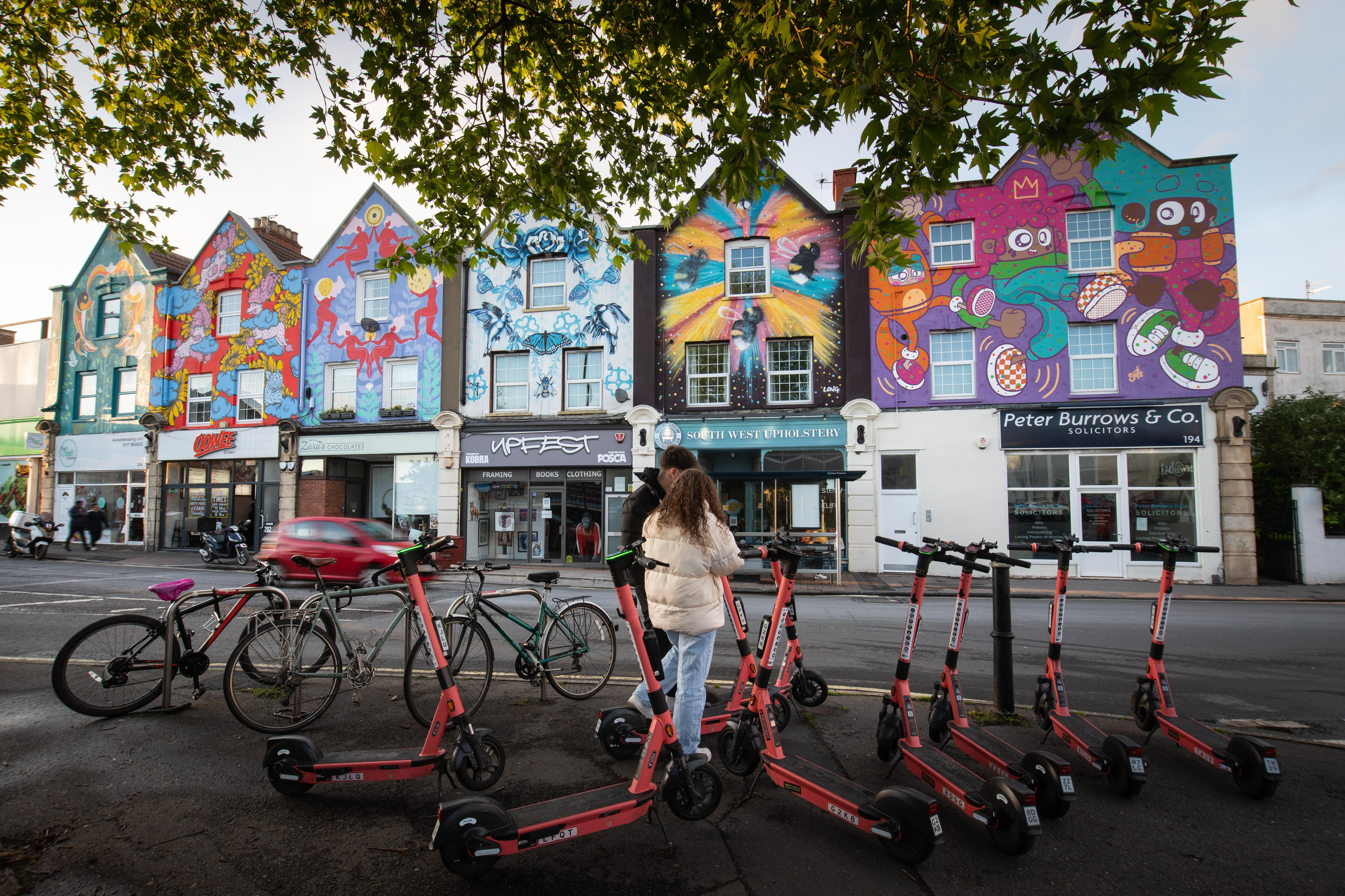 Two people standing by rental scooters in front of a row of houses painted on with colourful murals