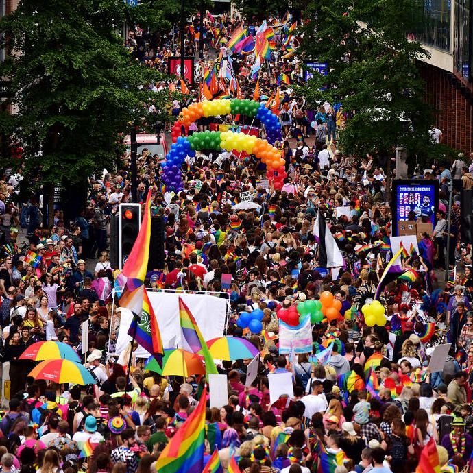 Multitud marchando en un desfile, con banderas arcoíris, paraguas y globos.