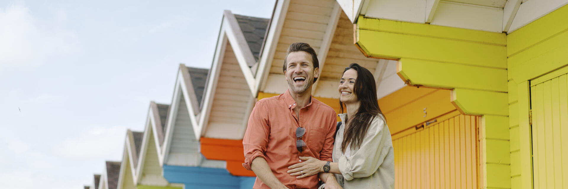 A man and woman laughing outside colourful beach huts