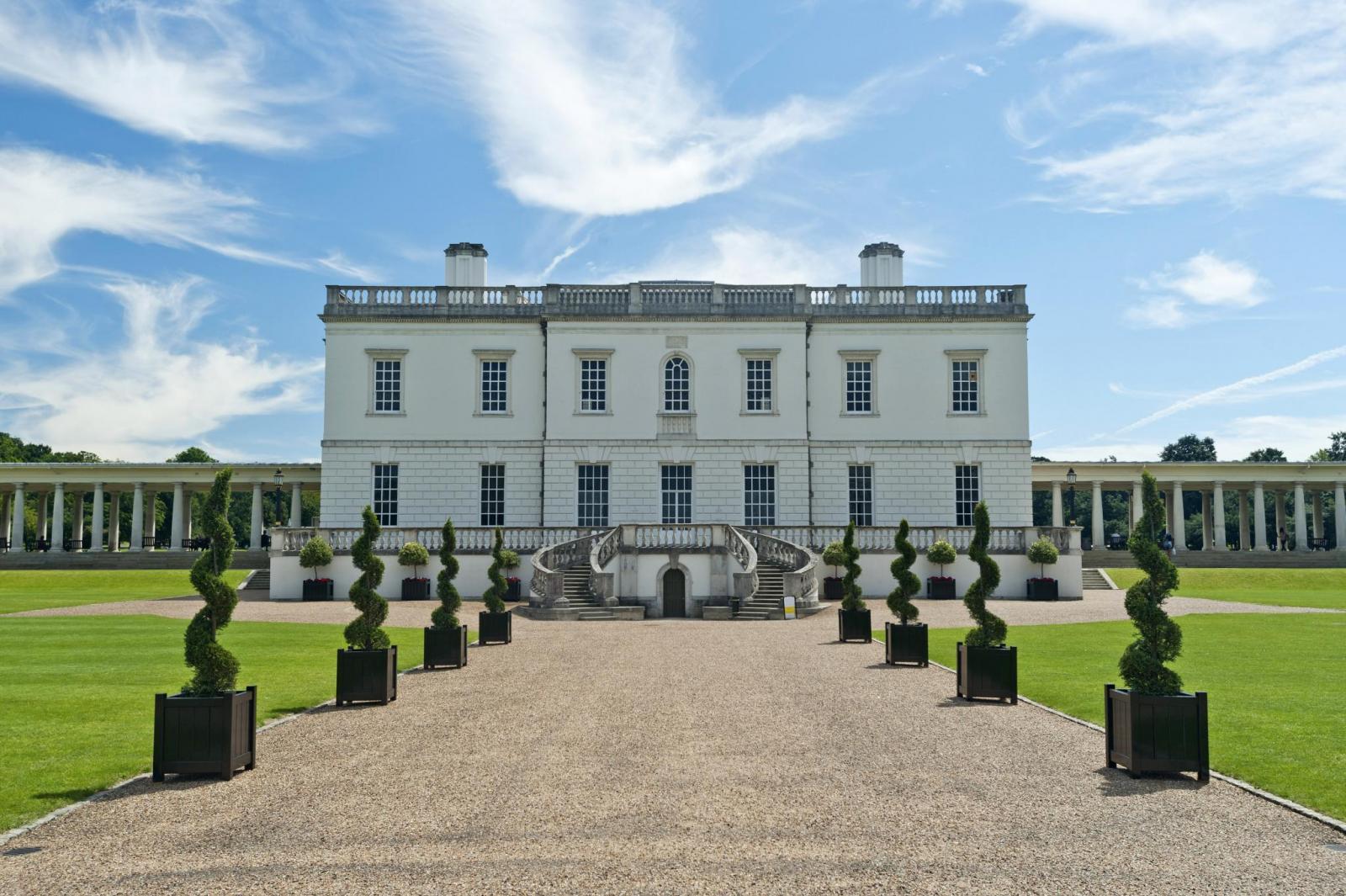 Grande maison blanche avec jardin, allée bordée d’arbres taillés, sous un ciel bleu et nuageux.