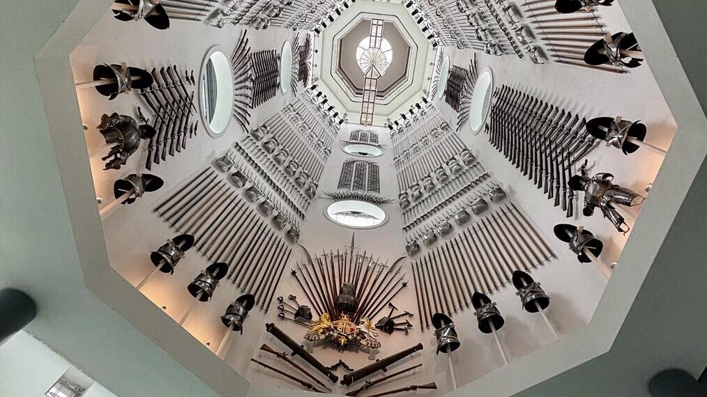 View up an octagonal stairwell with walls decorated by arranged armor, helmets, and weapons in a museum-like setting.