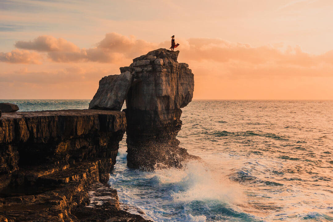Woman standing on a rock stack looking out to sea at sunset