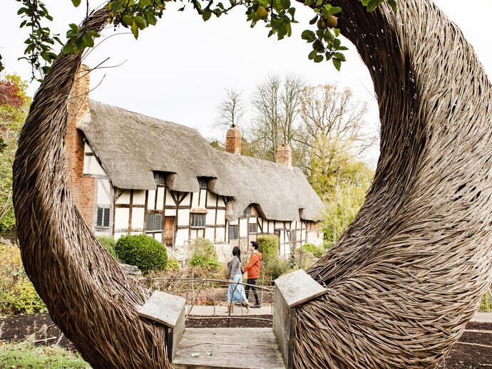 Couple walking near a cottage seen through a large willow crescent shaped sculpture in a garden