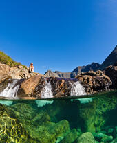 Clear stream with visible underwater rocks, small waterfall, rocky landscape and mountains under a blue sky. Person sits on rocks above the water.