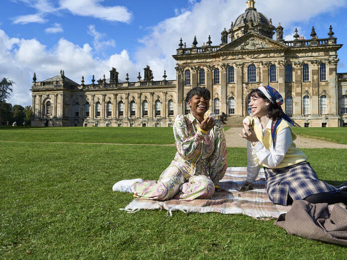 Two people enjoy a picnic on a blanket in front of a large historic building with ornate architecture on a sunny day.