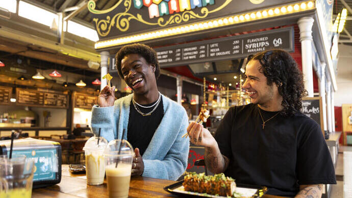 Dos personas sentadas en una mesa de un mercado cubierto, riendo y comiendo, con un letrero colorido de "MARKET" al fondo.