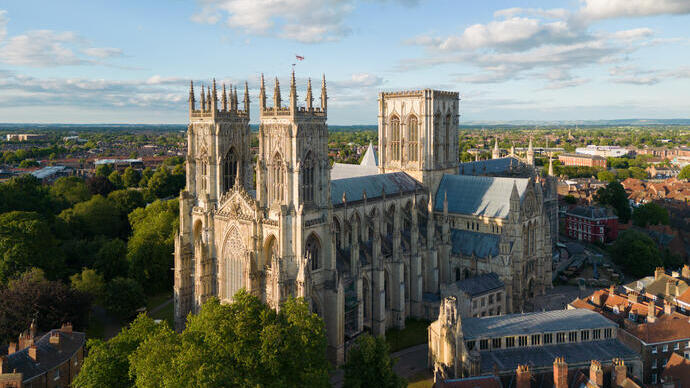 Large Gothic-style cathedral surrounded by trees and historic buildings under a partly cloudy sky, cityscape in background.