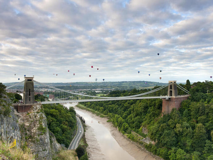 Hot air balloons floating above a river and a suspension bridge.