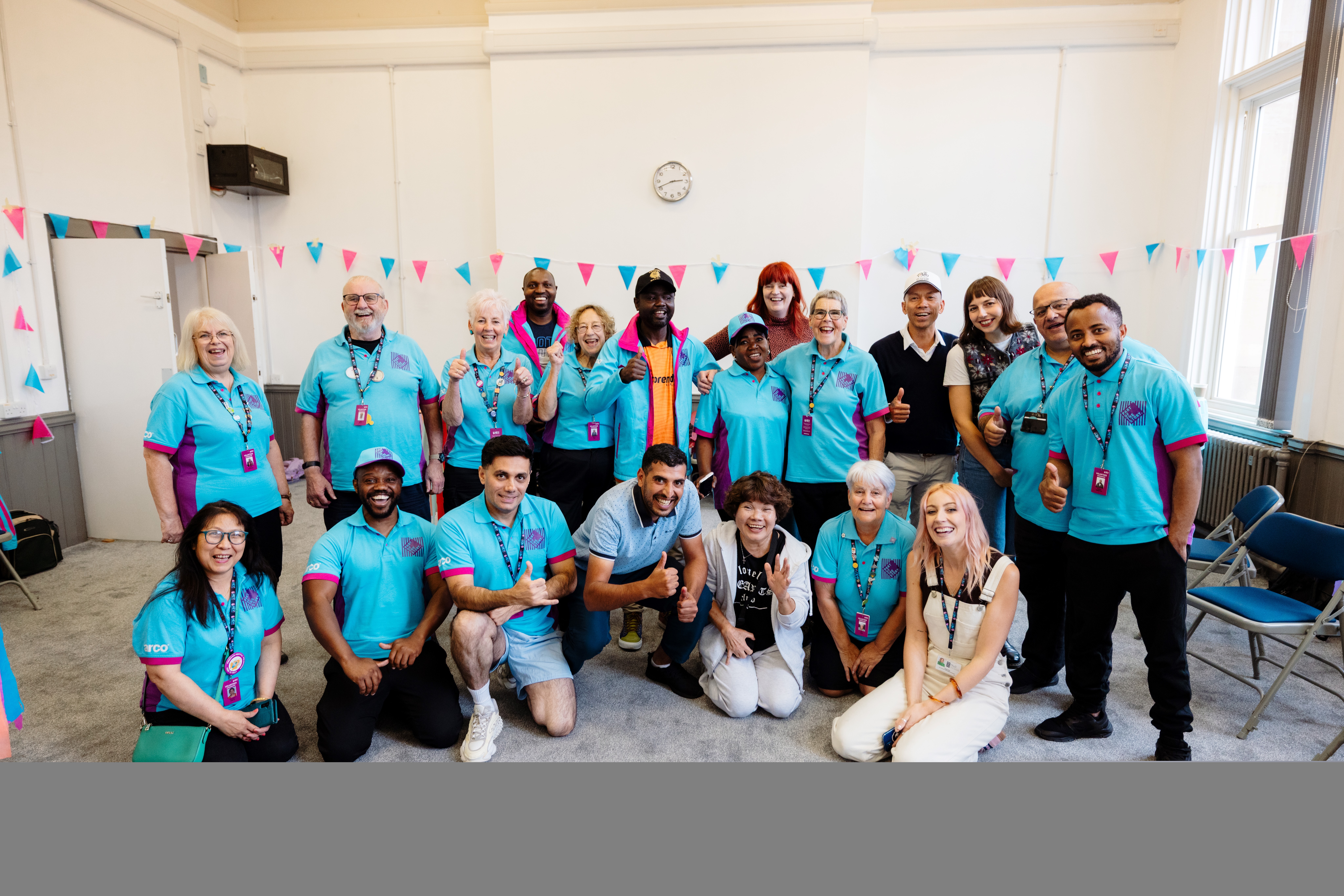 A diverse group of people in matching blue shirts gather in a decorated room, posing and smiling for a group photo, suggesting a team or community event.